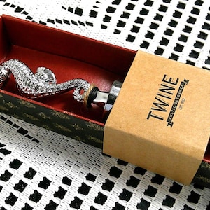May include: A decorative seahorse bottle stopper in a gift box. The stopper is silver-colored and shaped like a seahorse. The box is red inside with a black and gold patterned exterior. The box has a brown paper sleeve with the word "TWINE" printed on it.