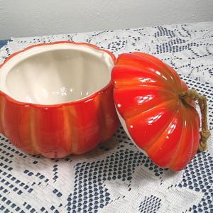 May include: A ceramic pumpkin-shaped serving bowl with a lid. The bowl is bright orange with darker orange shading and a white interior. The lid has a rope handle. The bowl is open, revealing the white interior. The bowl sits on a white lace tablecloth.