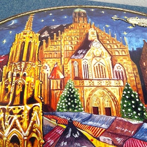May include: A decorative plate with a painted scene of a European city at night. The image depicts illuminated buildings, a starry sky, and a festive atmosphere. The plate has a gold trim around the edge.