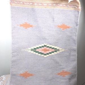 May include: A woven textile with a light purple background and geometric designs. The top section is brown with a patterned border. The central design is a diamond shape with green, pink, and white elements. Orange diamond shapes are scattered across the purple field.