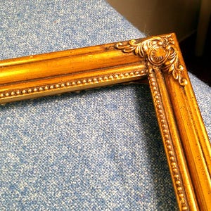 May include: Close-up of a section of a gold-colored picture frame. The frame has ornate detailing at the corners and along the edges, with a beaded inner border. The frame is resting on a blue textured surface.
