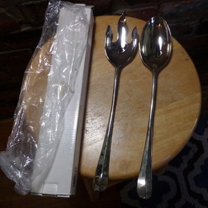 Op de afbeelding: Een set van twee zilveren serveerbestek, een lepel en een vork, in een doorzichtige plastic verpakking. Het bestek ligt op een houten oppervlak.