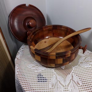 May include: A wooden salad bowl with a lid and a wooden spoon. The bowl is made of different types of wood, creating a checkered pattern. The lid is a solid piece of wood.