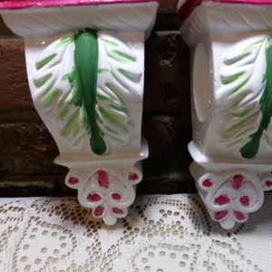 Puede incluir: Par de soportes de pared de cerámica blanca con detalles pintados en verde y rosa. Los soportes tienen un diseño de hojas y un borde inferior festoneado.