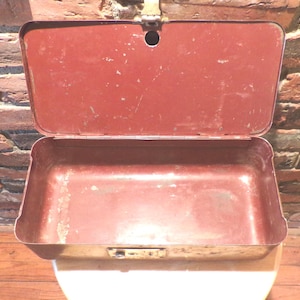 May include: Open, vintage red metal lunchbox with a hinged lid and a latch closure. The interior is empty, showing signs of wear. The lunchbox is set against a brick wall and wooden surface.