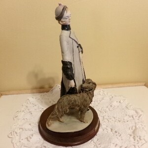 Pu&ograve; includere: Una statuetta in porcellana di una donna con un cappotto bianco e un cappello nero, che passeggia con un cane marrone. La statuetta &egrave; su una base di legno.