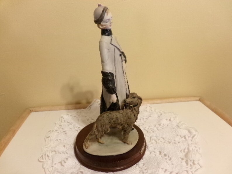 Pu&ograve; includere: Una statuetta in porcellana di una donna con un cappotto bianco e un cappello nero, che passeggia con un cane marrone. La statuetta &egrave; su una base di legno.