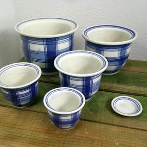 Peut inclure: Un ensemble de quatre cache-pots en céramique et une petite soucoupe assortie, tous dotés d'une base blanche avec un motif à carreaux bleus. Les cache-pots varient en taille, chacun avec un large rebord et un corps arrondi et lisse.