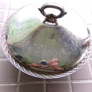 May include: A silver serving dish with a lid and a decorative handle. The dish has a raised edge and a floral pattern.