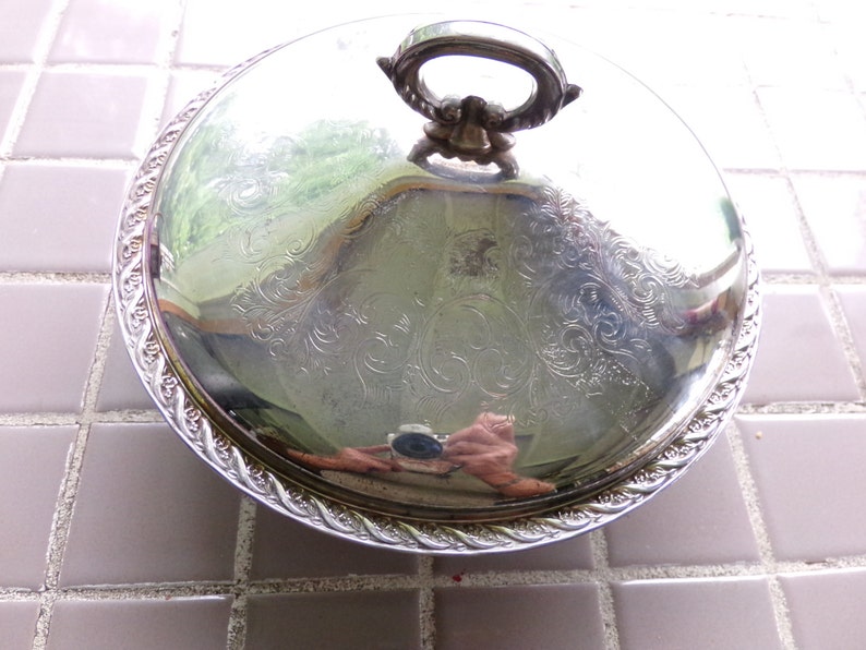 May include: A silver serving dish with a lid and a decorative handle. The dish has a raised edge and a floral pattern.