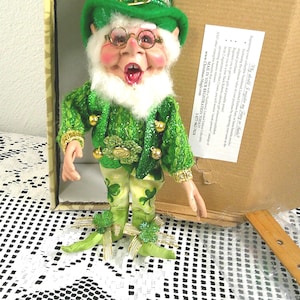 Puede incluir: Una muñeca de duende del Día de San Patricio. La muñeca lleva un sombrero, una chaqueta y pantalones verdes con detalles dorados y de tréboles. Tiene barba blanca, gafas y la boca abierta. La muñeca está en una caja de cartón.
