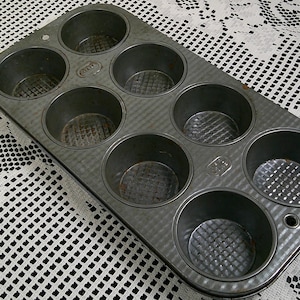 May include: A rectangular, metal muffin tin with eight circular molds. The tin is a dull silver color and sits on a white lace tablecloth. The molds have a textured, ridged pattern on the bottom.