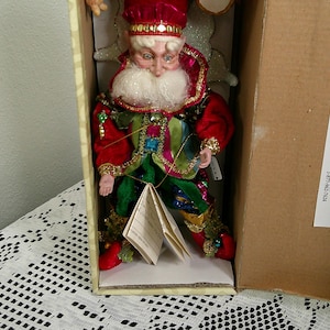 Könnte beinhalten: Dekorative Weihnachtselfenpuppe in einem Karton. Der Elf trägt einen roten Samthut und -mantel mit goldenen Verzierungen, eine grüne Weste und eine bunte Hose. Die Puppe hat einen weißen Bart und hält ein kleines Büchlein.
