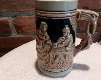 Puede incluir: Una jarra de cerveza de cerámica con un esmalte marrón y blanco. La jarra presenta un diseño en relieve de dos personas sentadas en una mesa. El asa tiene forma de rama de árbol.