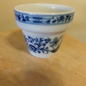 Puede incluir: Maceta de cerámica blanca con un diseño floral azul. La maceta tiene un borde ancho con un borde floral azul. El cuerpo de la maceta presenta un detallado patrón floral azul. La maceta está sobre una superficie de madera.