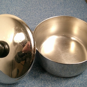 May include: A shiny, chrome-colored pot with a lid. The lid has a black handle and a green knob. The pot is open, revealing a clean, silver interior. The pot is likely used for cooking or food storage.