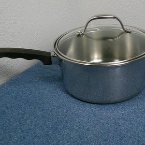 Może przedstawiać: Garnek ze stali nierdzewnej ze szklaną pokrywką i czarnym uchwytem. Garnek stoi na niebieskiej powierzchni. Uchwyt jest zakrzywiony i ergonomiczny.