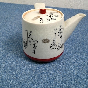 May include: A white ceramic teapot with a red base and lid accent. The teapot features black calligraphy and a spout. The lid has a small red knob. The teapot is on a blue surface.