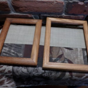 Puede incluir: Dos marcos de fotos de madera, uno marrón claro y otro marrón oscuro, se colocan uno al lado del otro sobre una superficie de tela estampada. Ambos marcos están vacíos y tienen un diseño rectangular simple.