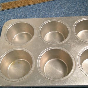 Pode incluir: Uma forma para muffins de seis cavidades feita de metal prateado. A forma tem formato retangular com bordas arredondadas e seis cavidades circulares. Um pequeno pedaço retangular de cortiça com borda metálica está ao fundo.