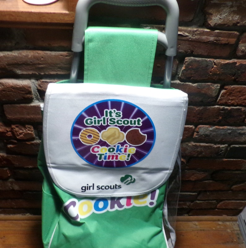 May include: A green and white rolling cart with a Girl Scout Cookie theme. The cart has a white panel with the text "It's Girl Scout Cookie Time!" and a logo with three cookies. The cart also has the text "girl scouts" and "Cookie!" printed on it.
