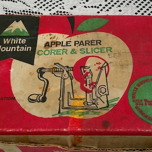 May include: A vintage red box for an apple parer, corer, and slicer. The box features a graphic of an apple with the words "Apple Parer Corer & Slicer." The brand name "White Mountain" is also visible.