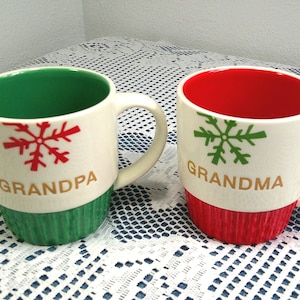 May include: Two ceramic mugs with white exteriors, green and red bases, and matching interiors. One mug has a red snowflake and the word "GRANDPA", the other has a green snowflake and the word "GRANDMA".
