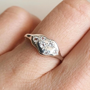May include: A silver ring with a unique, organic shape, set with several small, clear gemstones. The ring is worn on a finger, showcasing its design and sparkle. The ring's design is modern and elegant.
