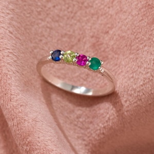 以下が含まれることがあります： 青、黄緑、マゼンタ、緑の4つのカラーストーンが付いたシルバーリング。 リングはピンク色の背景に配置され、石の鮮やかな色を際立たせています。