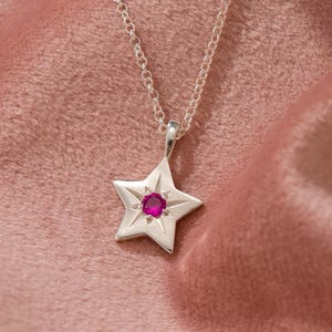 May include: A silver star pendant necklace with a small, round, red gemstone in the center. The star has five points and hangs from a delicate chain. The background is a soft, pink fabric.