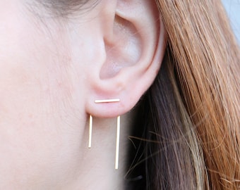 May include: Gold-colored bar earrings. The earrings feature a horizontal bar that sits on the earlobe, with a vertical bar hanging below. The earrings are simple and modern in design.