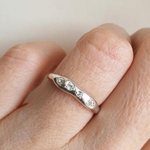 Può includere: Un anello d'argento dal design ondulato, incastonato con tre piccole gemme trasparenti. L'anello è indossato su un dito, mettendo in risalto il suo metallo lucido e le pietre scintillanti. Il design è semplice ma elegante.