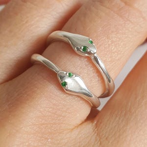 Puede incluir: Dos anillos de plata con forma de serpiente, cada uno con una banda de metal lisa y pulida que se envuelve alrededor del dedo. La cabeza de cada serpiente está detallada con dos pequeñas piedras verdes vibrantes como ojos, creando un diseño.