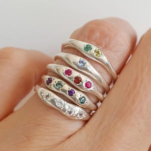 Puede incluir: Una pila de cinco anillos de plata, cada uno con una banda lisa y orgánica. Los anillos están engastados con varias piedras preciosas pequeñas, redondas y multicolores, incluyendo piedras transparentes, moradas, verde azulado y rojas.