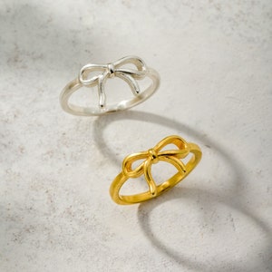 May include: Two rings, one silver and one gold, each with a bow design. The rings are displayed on a neutral, textured surface. The gold ring has a shiny finish, while the silver ring has a more matte appearance.