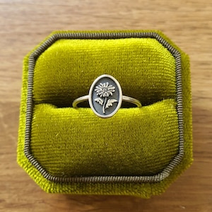 Pu&ograve; includere: Un anello d'argento con un design a forma di fiore in una scatola per anelli in velluto verde.