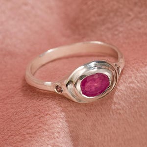 May include: A silver ring with a vibrant oval-shaped ruby gemstone. The ring's band is smooth and polished, with a decorative design around the ruby. The ring is set against a soft, pink background.