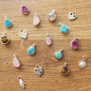 Op de afbeelding: Een verzameling van 17 zilveren bedelarmbanden met verschillende edelstenen, waaronder turquoise, tijgeroog, robijn en rozekwarts. De bedels worden getoond op een houten oppervlak.