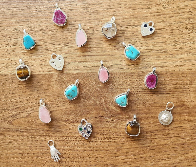 Op de afbeelding: Een verzameling van 17 zilveren bedelarmbanden met verschillende edelstenen, waaronder turquoise, tijgeroog, robijn en rozekwarts. De bedels worden getoond op een houten oppervlak.