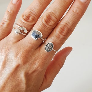 Pu&ograve; includere: Una mano con tre anelli d'argento. L'anello sul dito medio ha una pietra blu, l'anello sull'indice ha un design arcobaleno e l'anello sull'anulare ha un design floreale.