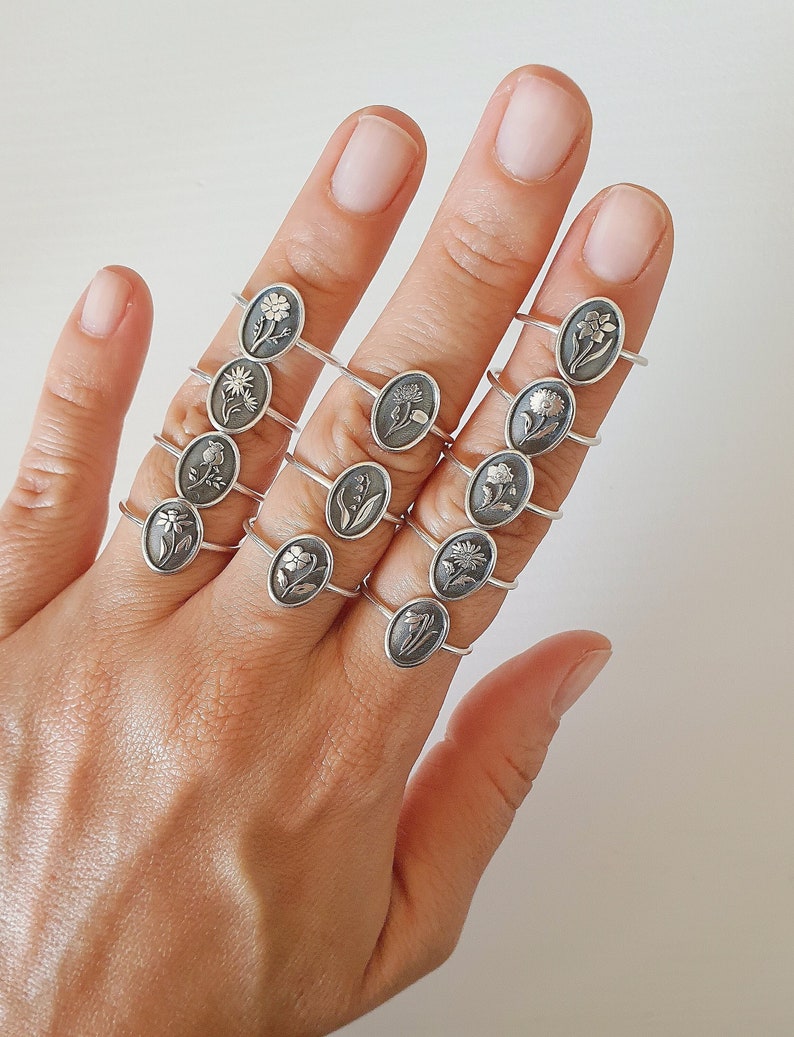 Pu&ograve; includere: Un set di anelli d'argento con diversi disegni di fiori. Ogni anello presenta un motivo floreale unico, tra cui una margherita, un tulipano, un narciso e un giglio. Gli anelli sono impilati su una mano, mostrando i dettagli intricati di ogni design.