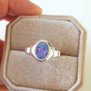 May include: A silver ring featuring an oval opal gemstone. The opal exhibits a colourful play of blues, purples, and pinks. The ring is set in a polished silver band. The ring is displayed in a beige velvet ring box.
