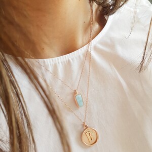 May include: Two gold necklaces with pendants. The top necklace has a light blue rectangular pendant. The bottom necklace has a round pendant with the letter "B" engraved on it.