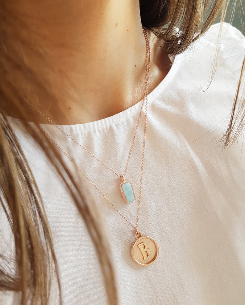 May include: Two gold necklaces with pendants. The top necklace has a light blue rectangular pendant. The bottom necklace has a round pendant with the letter "B" engraved on it.