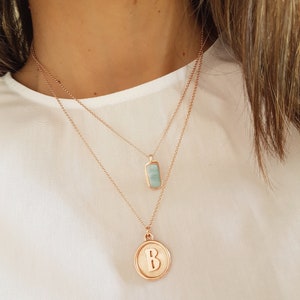 May include: Two gold necklaces with pendants. The top necklace has a light blue rectangular pendant. The bottom necklace has a round pendant with the letter "B" in gold.