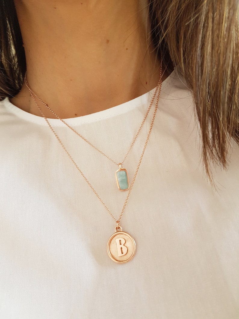 May include: Two gold necklaces with pendants. The top necklace has a light blue rectangular pendant. The bottom necklace has a round pendant with the letter "B" in gold.