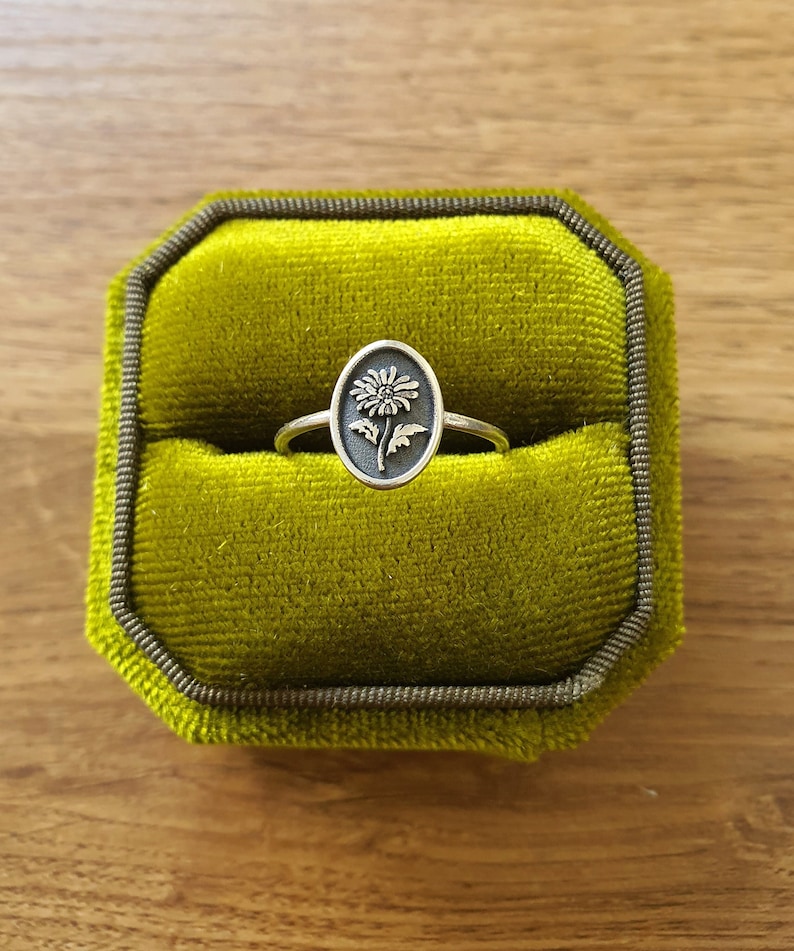 Pu&ograve; includere: Un anello d'argento con un design a forma di fiore in una scatola per anelli in velluto verde.