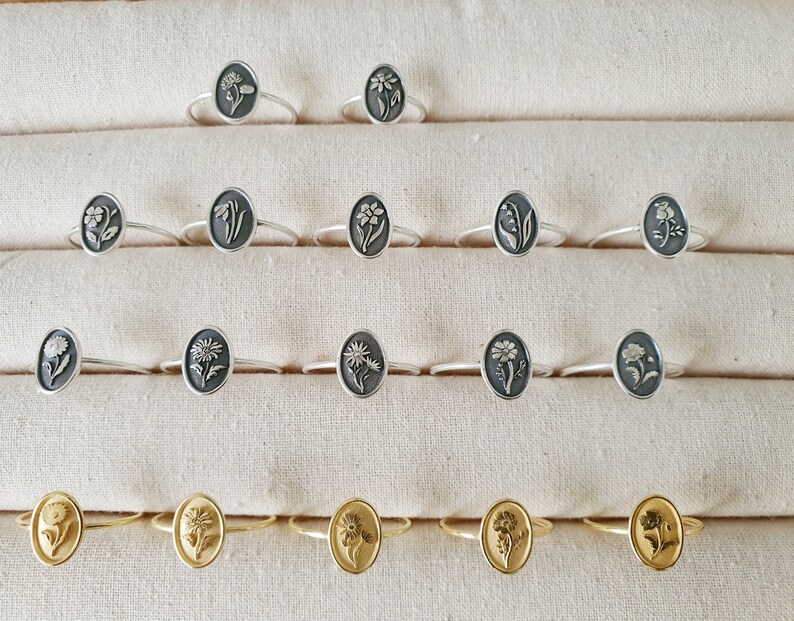Pu&ograve; includere: Una esposizione di anelli in argento e oro con motivi floreali. Gli anelli sono disposti in file su uno sfondo di tessuto bianco.