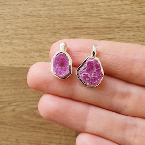 Puede incluir: Dos colgantes de plata con piedras de rubí rosa. Las piedras son ásperas y tienen un aspecto natural y sin pulir.