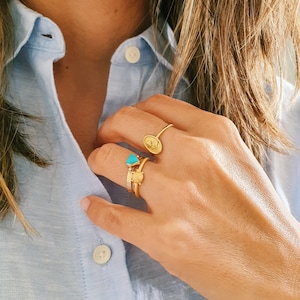 Pu&ograve; includere: Una mano con tre anelli d'oro. Gli anelli presentano un cuore turchese, un piccolo cuore d'oro e un ovale pi&ugrave; grande con un design in rilievo.